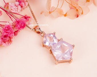 May include: A rose gold necklace with a pendant featuring two light pink, geometric-cut gemstones. The pendant is suspended from a delicate chain. The background includes pink flowers and the text "azgemsnjewelry".