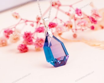 Op de afbeelding: Een zilveren ketting met een grote, facetgeslepen hanger. De hanger heeft een druppelvorm met een kleurovergang van blauw naar paars. De ketting wordt gepresenteerd tegen een zachte, vage bloemenachtergrond.