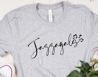 May include: A gray t-shirt with the text "Jazzagals" and musical notes in black lettering. The shirt is laid out on a white wooden surface with a pair of white sneakers, a pair of denim shorts, and a sprig of greenery.