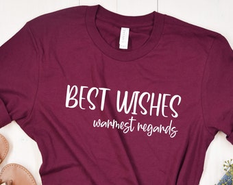 May include: A maroon t-shirt with the text "BEST WISHES warmest regards" in white script. The shirt is paired with denim shorts, gold sandals, and greenery, all arranged on a white wooden surface. A casual, lifestyle shot.