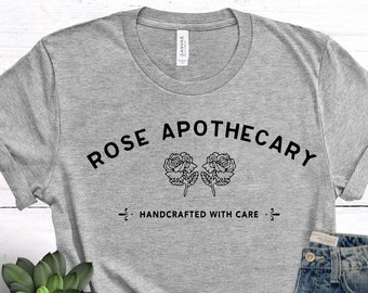 May include: A gray t-shirt with the text "ROSE APOTHECARY" and two roses printed on it. The text "HANDCRAFTED WITH CARE" is printed below the roses. The t-shirt is paired with blue jeans and pink sneakers.