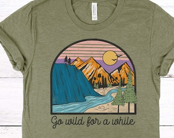 Puede incluir: Una camiseta verde con un gráfico de una cordillera, un río y una puesta de sol. El texto "Go wild for a while" está impreso debajo del gráfico. La camiseta es de color verde oliva jaspeado.