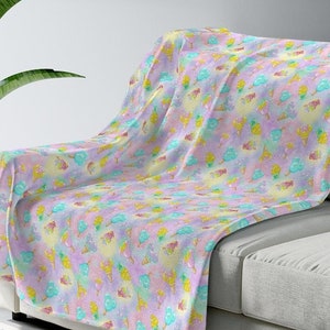 May include: A soft pink, yellow, and blue fleece throw blanket with a repeating pattern of ice cream cones. The blanket is draped over a light gray couch.