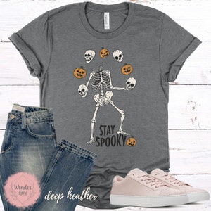 K&ouml;nnte beinhalten: Ein graues T-Shirt mit einem Skelett, das K&uuml;rbisse und Sch&auml;del jongliert. Der Text "Stay Spooky" ist unter dem Skelett aufgedruckt.