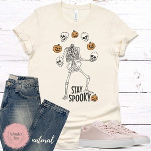 K&ouml;nnte beinhalten: Ein beiges T-Shirt mit einem Skelett, das K&uuml;rbisse und Sch&auml;del jongliert. Der Text "Stay Spooky" ist auf dem T-Shirt aufgedruckt.