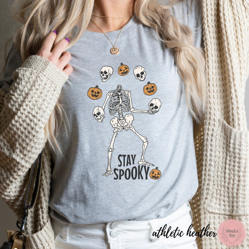 K&ouml;nnte beinhalten: Ein graues T-Shirt mit einem Skelett, das K&uuml;rbisse und Sch&auml;del jongliert. Der Text "Stay Spooky" ist unter dem Skelett gedruckt. Das Shirt ist mit "athletic heather" gekennzeichnet.