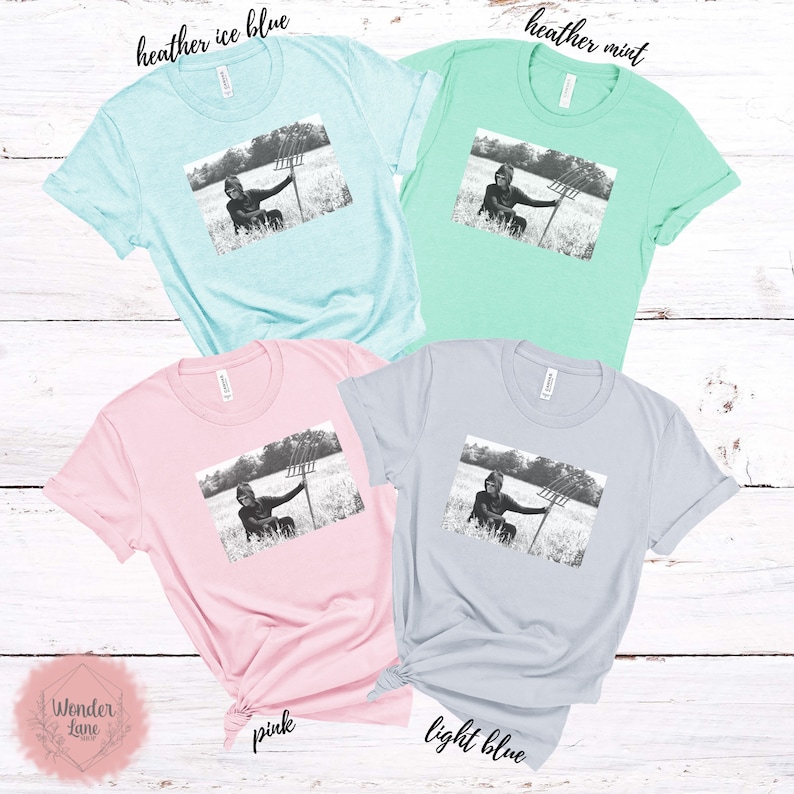May include: Four t-shirts in different colors, each featuring a black and white image of a person holding a pitchfork in a field. The t-shirts are heather ice blue, heather mint, pink, and light blue.