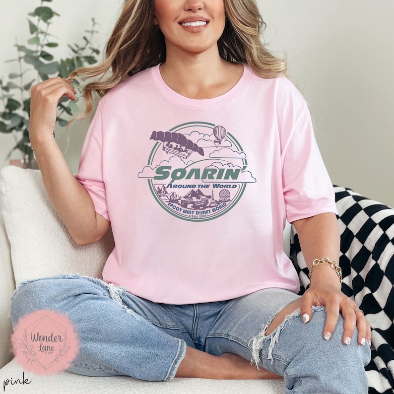 May include: A pink t-shirt with a graphic design of a ride called "Soarin' Around the World" at Epcot Walt Disney World. The graphic features a hot air balloon, a ride vehicle, and a pyramid.