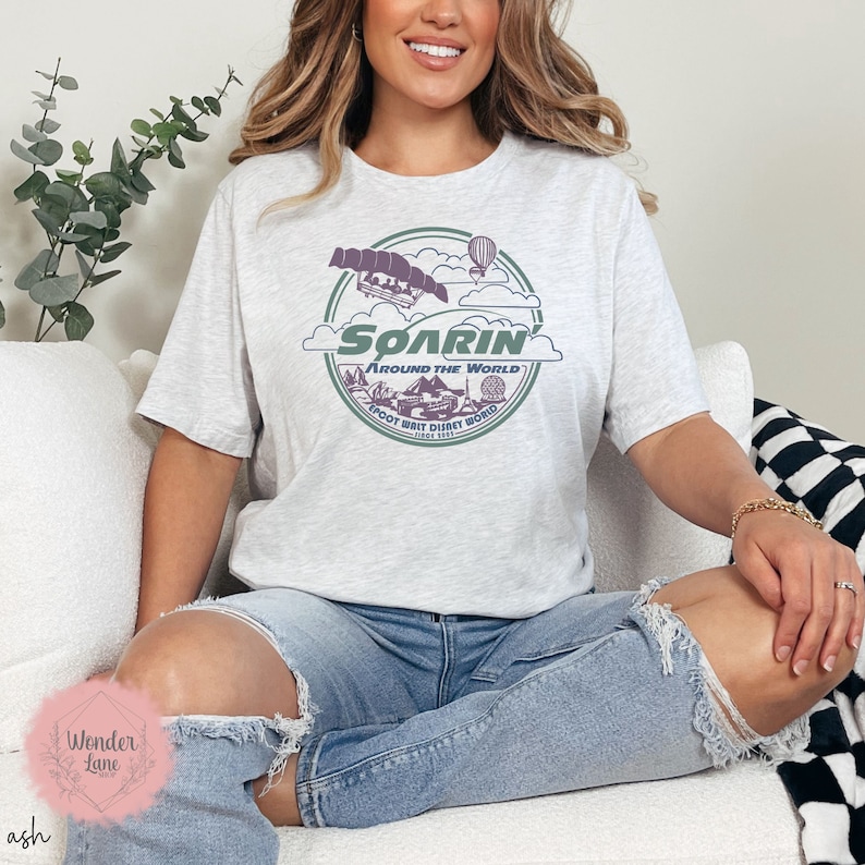 May include: A gray t-shirt with a graphic of the Soarin' Around the World ride at Epcot. The graphic features a purple and green color scheme and includes the text "Soarin' Around the World Epcot Walt Disney World Since 1982".