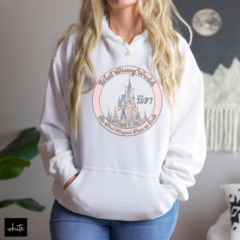 Puede incluir: Sudadera blanca con capucha con un gr&aacute;fico circular rosa que presenta un dibujo animado del castillo de Cenicienta con el texto "Walt Disney World since 1971 The Most Magical Place on Earth".