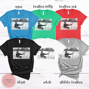 May include: A black and white photo of a person holding a pitchfork in a field. The photo is printed on a variety of t-shirts in different colors, including aqua, heather kelly, heather red, black, white, and athletic heather.
