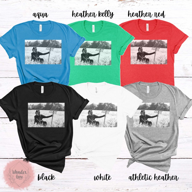 May include: A black and white photo of a person holding a pitchfork in a field. The photo is printed on a variety of t-shirts in different colors, including aqua, heather kelly, heather red, black, white, and athletic heather.