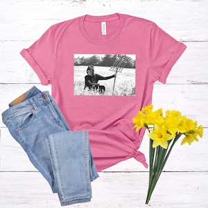 May include: A pink t-shirt with a black and white image of a person wearing a hooded sweatshirt and holding a pitchfork in a field.