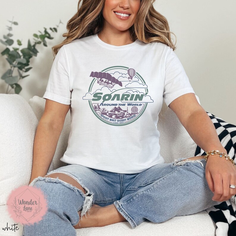May include: White t-shirt with a graphic of the Soarin' Around the World ride at Epcot Walt Disney World. The graphic features a purple and green color scheme with a hot air balloon and a ride vehicle.
