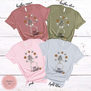 K&ouml;nnte beinhalten: Vier T-Shirts mit einem Skelett, das K&uuml;rbisse und Sch&auml;del jongliert. Die Shirts sind in Heather Mauve, Heather Olive, Pink und Hellblau. Der Text auf den Shirts lautet "Stay Spooky".