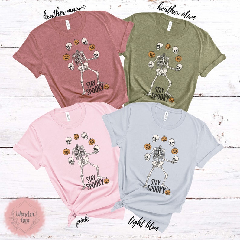 K&ouml;nnte beinhalten: Vier T-Shirts mit einem Skelett, das K&uuml;rbisse und Sch&auml;del jongliert. Die Shirts sind in Heather Mauve, Heather Olive, Pink und Hellblau. Der Text auf den Shirts lautet "Stay Spooky".