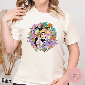 May include: A white t-shirt with a colorful floral design featuring the faces of Disney villains. The villains include Maleficent, Cruella de Vil, Ursula, and the Evil Queen. The text "Wonder Lane" is visible in the bottom right corner.