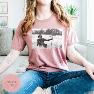 May include: A black and white photo of a person wearing a hooded sweatshirt and holding a pitchfork in a field. The photo is printed on a pink t-shirt.