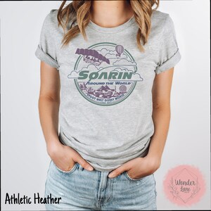 May include: A gray t-shirt with a vintage-style graphic of the Soarin' Around the World ride at Epcot. The graphic features a purple and green color scheme and includes the text "Soarin' Around the World" and "Epcot Walt Disney World Since 1982".