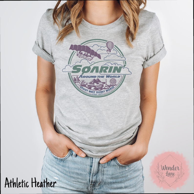May include: A gray t-shirt with a vintage-style graphic of the Soarin' Around the World ride at Epcot. The graphic features a purple and green color scheme and includes the text "Soarin' Around the World" and "Epcot Walt Disney World Since 1982".