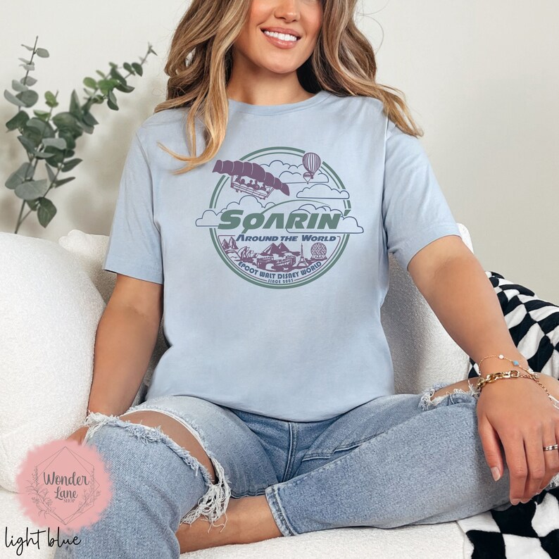 May include: Light blue t-shirt with a graphic of the Soarin' ride at Epcot, featuring a hot air balloon, a hang glider, and the text "Soarin' Around the World Epcot Walt Disney World Since 1982".
