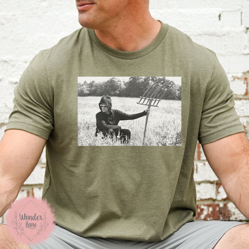 May include: A green t-shirt with a black and white photo of a person wearing sunglasses and a hooded sweatshirt holding a pitchfork in a field.