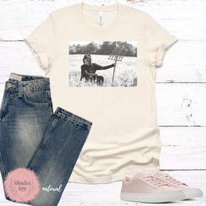 May include: A black and white photo of a person wearing a hooded sweatshirt and holding a pitchfork in a field. The photo is printed on a cream colored t-shirt.