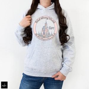Puede incluir: Una sudadera gris con un gr&aacute;fico circular rosa que presenta una ilustraci&oacute;n de dibujos animados del castillo de Cenicienta. El texto en el gr&aacute;fico dice "Walt Disney World since 1971 The Most Magical Place on Earth".