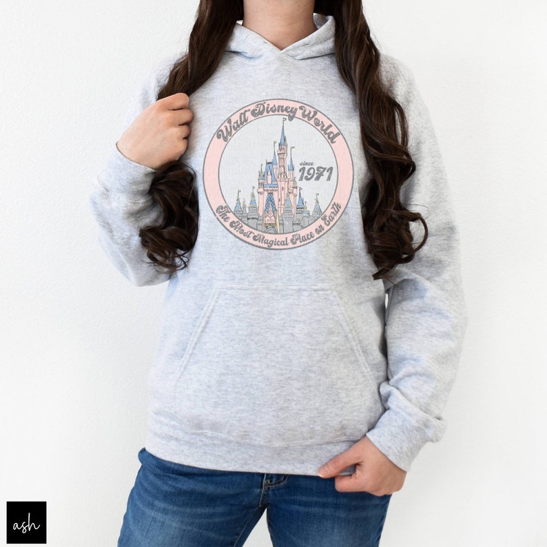 Puede incluir: Una sudadera gris con un gr&aacute;fico circular rosa que presenta una ilustraci&oacute;n de dibujos animados del castillo de Cenicienta. El texto en el gr&aacute;fico dice "Walt Disney World since 1971 The Most Magical Place on Earth".