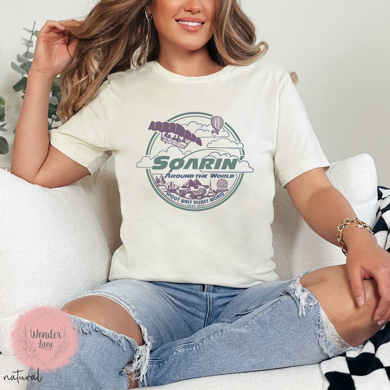 May include: A white t-shirt with a green and purple graphic of the Soarin' Around the World ride at Epcot. The graphic features a hot air balloon, a ride vehicle, and the text "Soarin' Around the World".