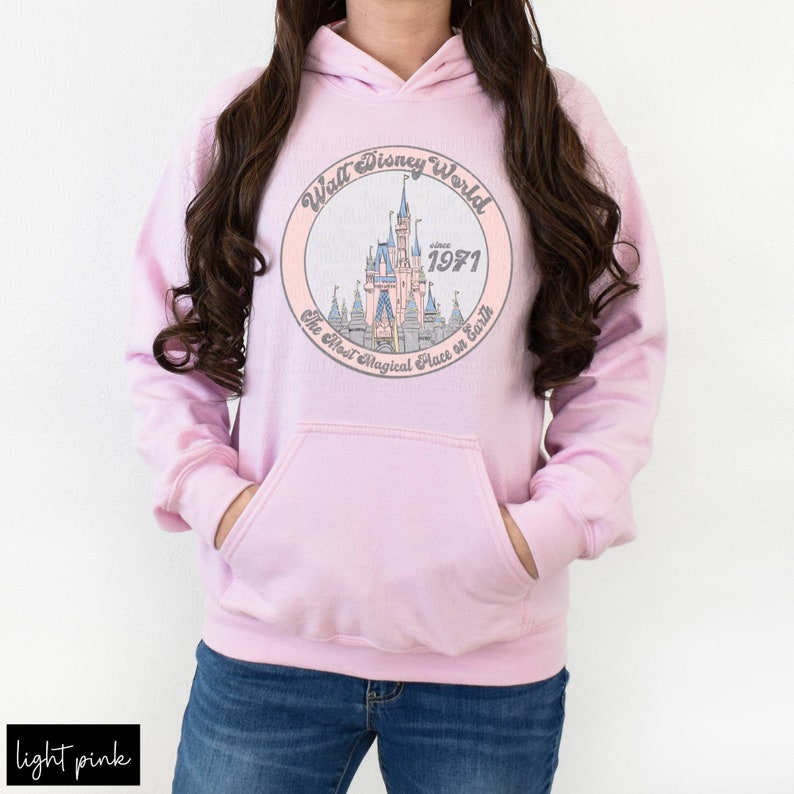 Puede incluir: Una sudadera con capucha rosa claro con un gr&aacute;fico circular blanco que presenta un dibujo animado del castillo de Cenicienta con el texto "Walt Disney World since 1971 The Most Magical Place on Earth".