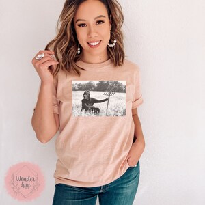 May include: A black and white photo of a person sitting in a field holding a pitchfork. The photo is printed on a light pink t-shirt.