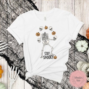 K&ouml;nnte beinhalten: Wei&szlig;es T-Shirt mit einem Skelett, das K&uuml;rbisse jongliert, und dem Text "Stay Spooky".
