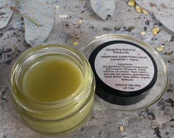 May include: A small glass jar of green salve with a black lid. The lid has a label that reads "Jacquie's Natural Products Handmade Everything Salve Dandelion + Monoi" and lists the ingredients: "Dry Dandelion Flowers (taraxacum officinaleni) infused in Monoi Tahitian Oil (monoi tiare tahiti). Avocado Oil (persea, gratissima). Monoi Butter, Shea Butter, Organic Beeswax" and "External Use Only wt 1 oz".