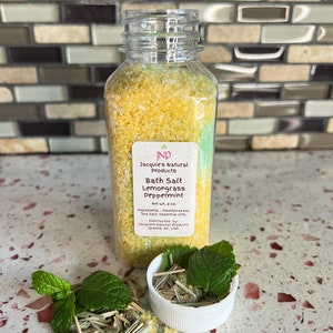 Puede incluir: Una botella de plástico transparente llena de sales de baño amarillas. La etiqueta dice "INP Jacquie's Natural Products Bath Salt Lemongrass Peppermint Net Wt. 8 oz Ingredients - Mediterranean Sea Salt, Essential Oils, Distributed by Jacquie's Natural Products Queens, NY, USA". Hay ramitas de menta fresca y hierba de limón seca esparcidas alrededor de la botella.