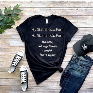 Może przedstawiać: Czarny t-shirt z białym tekstem: "Ho: Statistics = Fun, HA: Statistics ≠ Fun, The only null hypothesis I would fail to reject." Zawiera czarne trampki, niebieskie dżinsy i czapkę Adidas.