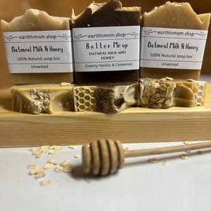 May include: Three bars of handmade soap with labels that read "Oatmeal Milk & Honey", "Butter Me Up Oatmeal Milk and Honey Creamy Vanilla & Cinnamon", and "Oatmeal Milk & Honey". The soap bars are arranged on a wooden surface with a honey dipper and oats scattered around them.