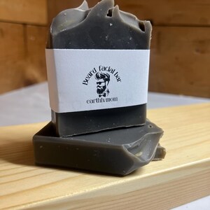 May include: Two bars of dark gray soap stacked on top of each other. The top bar has a white label with the text "Beard facial bar" and "earthlymom" printed in black.
