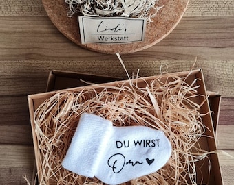 Könnte beinhalten: Ein Geschenkset mit einer weißen Babysocke mit dem Aufdruck "DU WIRST Oma" und einem Herzsymbol, präsentiert in einer Box mit Stroh. Im Hintergrund ein Blumenarrangement mit weißen Rosen und Trockenblumen.