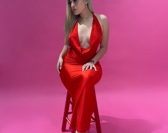 Puede incluir: Un vestido rojo con cuello halter y escote pronunciado. El vestido es largo hasta el suelo y se combina con tacones rojos. La persona está sentada en un taburete rojo sobre un fondo rosa. El vestido tiene un diseño elegante.