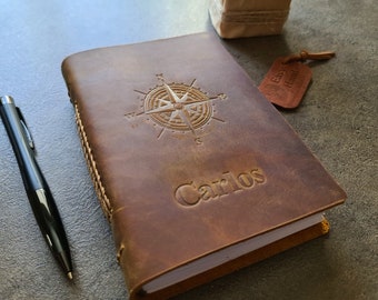 May include: A brown leather journal with a compass design and the name "Carlos" embossed on the front. The journal is closed and has a white paper insert.