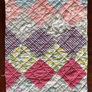 May include: A vintage quilt with a patchwork design. The quilt features a variety of fabrics in shades of blue, pink, red, and yellow. The pattern includes diamond and zigzag shapes, with a quilted texture. The quilt is likely handmade and shows signs of wear.