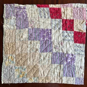 May include: A patchwork quilt with a variety of fabrics in cream, purple, red, and tan. The quilt is made of squares and rectangles, with a quilted pattern throughout. The quilt appears to be handmade and vintage.