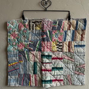 May include: A patchwork quilt with a variety of fabric patterns and colors, including floral, striped, and geometric designs. The quilt is displayed on a black metal hanger against a neutral wall. The quilt's colors include pink, blue, green, and white.