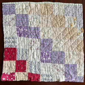 May include: A patchwork quilt with a variety of fabrics in shades of red, purple, cream, and tan. The quilt is made of squares and rectangles, with a light green border. The stitching is visible, creating a textured effect.