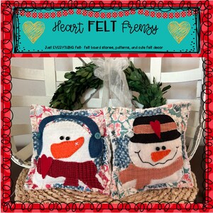 Puede incluir: Dos cojines decorativos de fieltro con motivos invernales. Uno representa un muñeco de nieve con sombrero negro y nariz naranja. El otro muestra un pingüino con orejeras y bufanda roja. Los cojines están sobre una cesta tejida.