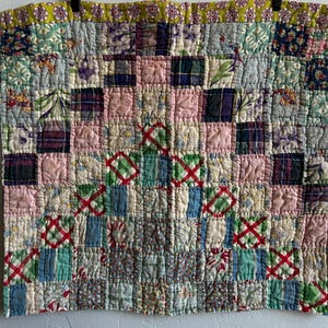 May include: A patchwork quilt with a variety of fabric squares in shades of pink, blue, green, and purple. The quilt features a repeating pattern of squares and diagonal red cross designs. The top edge has a floral patterned border.