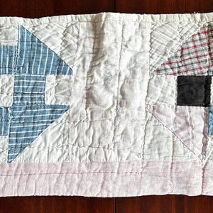May include: A rectangular, quilted textile featuring two geometric designs. One design is a blue and white diamond shape, and the other is a patchwork of red, black, and blue squares and rectangles. The background is white with a pink border.