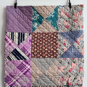 May include: A patchwork quilt with a variety of patterned fabrics in shades of purple, brown, blue, and gray. The quilt is hung on a black hanger against a white wall. The quilt is made of squares and triangles.