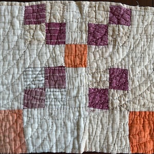 May include: Handmade quilt with a patchwork design. The quilt features a cream-colored background with squares of orange and purple. The quilt is made of soft fabric and has a quilted texture.